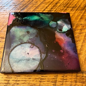 May include: A square ceramic tile with a black background and abstract art in shades of purple, pink, green, and blue. The art resembles a galaxy with swirling colours and white dots.