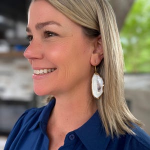 May include: A woman wearing gold-toned earrings. The earrings are large, irregular ovals with a white and gray interior and a gold-colored border. The woman is wearing a dark blue collared shirt.