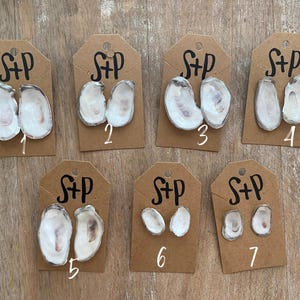 May include: Seven pairs of oyster shell earrings on brown cards with the text "S+P" written on each card. The cards are numbered 1 through 7.