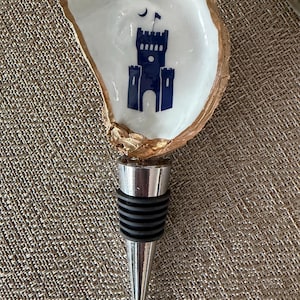 May include: A decorative bottle stopper featuring a white oyster shell with a gold rim. The shell is adorned with a blue castle design. The stopper has a silver metal base and a black rubber seal.