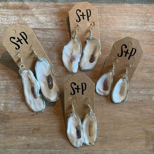 May include: Three pairs of oyster shell earrings on display cards with the text "S+P" on each card. The earrings are made from natural oyster shells with a white and brown color.
