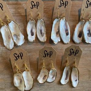 May include: Six pairs of gold oyster shell earrings with a white and brown interior. Each pair is displayed on a small card with the text "S+P" printed on it.