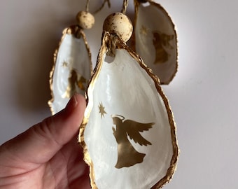 May include: Three white oyster shell ornaments with gold trim and a gold angel silhouette design. Each ornament has a small gold star and is strung together with twine and wooden beads.