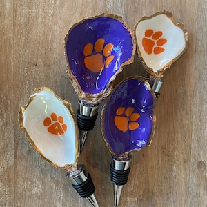 May include: Four decorative bottle stoppers. Each stopper features a painted oyster shell with a gold rim. Two shells are purple with orange paw prints, and two are white with orange paw prints. The stoppers have silver and black accents.