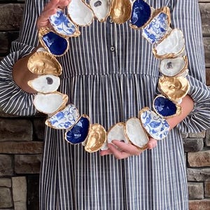 May include: A circular wreath constructed from individual oyster shells. The shells feature a variety of finishes, including solid gold, solid navy blue, plain white, and white with blue floral patterns, all arranged in a continuous ring formation.