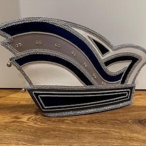Könnte beinhalten: Ein dekorativer Karnevalshut mit einem mehrschichtigen Design. Der Hut hat Abschnitte in Marineblau, Grau, Weiß und Schwarz, die mit silbernen Zierleisten umrandet sind. Kleine silberne Glöckchen und Strasssteine ​​verleihen dem Kopfschmuck Details.