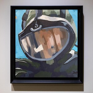 May include: An oil painting in a black frame depicts a close-up of a pilot's helmet with a reflective visor. The helmet is painted in shades of green, brown, and gray, set against a blue sky background. The artwork is signed in the lower right corner.