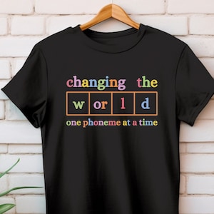 May include: Black t-shirt with the text "changing the world one phoneme at a time" in colorful letters. The word "world" is broken up into individual squares with each letter in a different color.