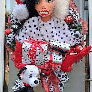 May include: A Christmas wreath with a Cruella de Vil figure wearing a Santa hat and a dalmatian-patterned coat. The wreath includes wrapped gifts, red ribbons, and a dalmatian dog. Red jingle bells hang at the bottom, creating a festive holiday theme.