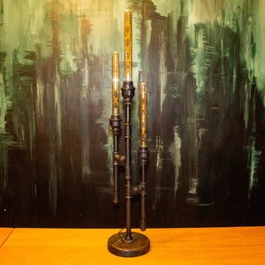 K&ouml;nnte beinhalten: Eine schwarze Tischlampe aus Metall mit vier Gl&uuml;hbirnen. Die Lampe hat ein Steampunk-Design mit Rohren und Zahnr&auml;dern. Die Lampe steht auf einem Holztisch.