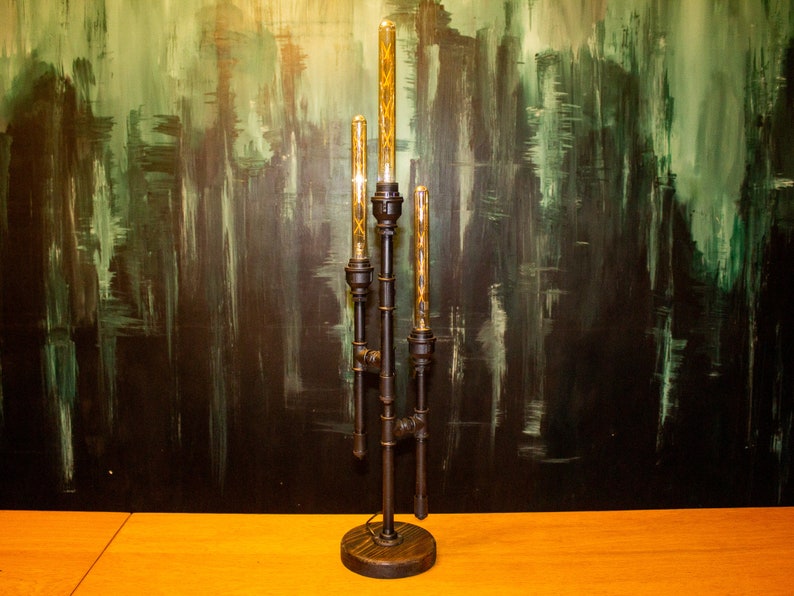 K&ouml;nnte beinhalten: Eine schwarze Tischlampe aus Metall mit vier Gl&uuml;hbirnen. Die Lampe hat ein Steampunk-Design mit Rohren und Zahnr&auml;dern. Die Lampe steht auf einem Holztisch.