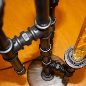 K&ouml;nnte beinhalten: Eine schwarze Metall-Rohrlampe mit einer durchsichtigen Gl&uuml;hbirne und einem Holzsockel. Die Lampe ist mit mehreren Rohrverbindungen zusammengesetzt und hat einen Vintage-Industriestyle.
