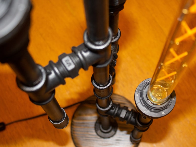 K&ouml;nnte beinhalten: Eine schwarze Metall-Rohrlampe mit einer durchsichtigen Gl&uuml;hbirne und einem Holzsockel. Die Lampe ist mit mehreren Rohrverbindungen zusammengesetzt und hat einen Vintage-Industriestyle.
