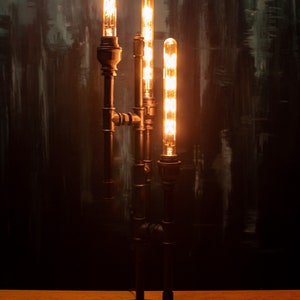 K&ouml;nnte beinhalten: Eine Steampunk-Stehlampe mit vier Edison-Gl&uuml;hbirnen. Die Lampe besteht aus schwarzen Metallrohren und hat einen Holzsockel.