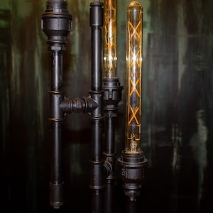 K&ouml;nnte beinhalten: Eine schwarze Metallrohrlampe mit drei Edison-Gl&uuml;hbirnenleuchten. Die Gl&uuml;hbirnen sind aus klarem Glas mit einem Filamentdesign.
