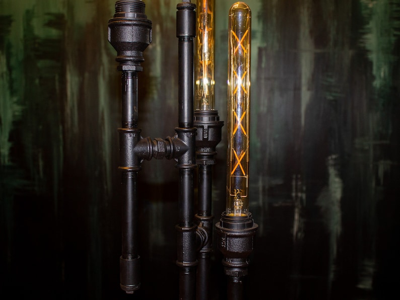 K&ouml;nnte beinhalten: Eine schwarze Metallrohrlampe mit drei Edison-Gl&uuml;hbirnenleuchten. Die Gl&uuml;hbirnen sind aus klarem Glas mit einem Filamentdesign.