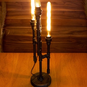 K&ouml;nnte beinhalten: Eine schwarze Tischlampe aus Metall mit drei Edison-Gl&uuml;hbirnen. Die Lampe hat ein rustikales Industriedesign und steht auf einem Holztisch.