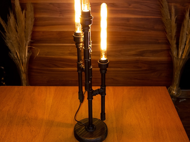 K&ouml;nnte beinhalten: Eine schwarze Tischlampe aus Metall mit drei Edison-Gl&uuml;hbirnen. Die Lampe hat ein rustikales Industriedesign und steht auf einem Holztisch.