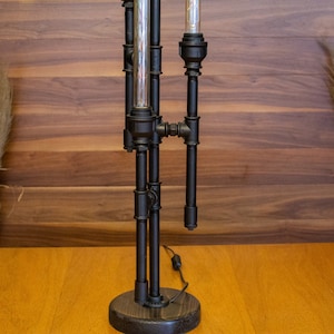 K&ouml;nnte beinhalten: Eine schwarze Tischlampe aus Metall mit einem einzigartigen Industriedesign. Die Lampe hat drei Gl&uuml;hbirnen und ein schwarzes Kabel.