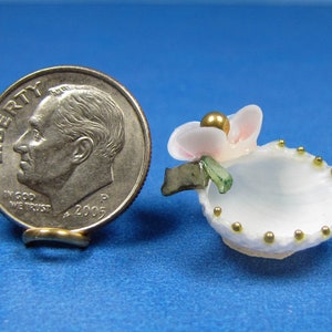 May include: A small white seashell with a pink flower on top. The shell is decorated with gold beads around the rim. The flower has a gold bead in the center.