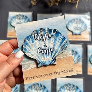 Może przedstawiać: Magnes w kształcie muszli z niebiesko-białymi paskami akwarelowymi, z imionami "Elisha & Angie" i datą "10.11.2026". Magnes znajduje się na kartce ze sceną plażową i tekstem "Thank you celebrating with us!"