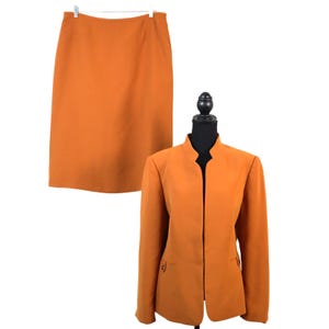 May include: A two-piece orange suit set. The set includes a knee-length skirt and a collared blazer with two small pockets. The blazer is displayed on a black mannequin. The skirt is hanging from a white hanger.