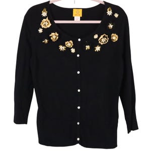 May include: Black cardigan with a scoop neck and long sleeves. The sweater features decorative gold and white floral embellishments around the neckline and down the front. It has white buttons and is displayed on a pink hanger.