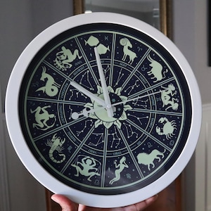 May include: A white-framed wall clock featuring a dark blue face with zodiac constellations and symbols. The clock hands are white, and the center has a sun design. The clock is approximately 12 inches in diameter.
