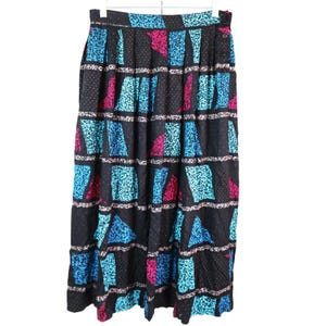 May include: A black midi skirt featuring a repeating geometric pattern of teal and magenta shapes interspersed with horizontal bands of white and black speckled detail. The skirt has a high waistband with a side button closure and pleated fabric.