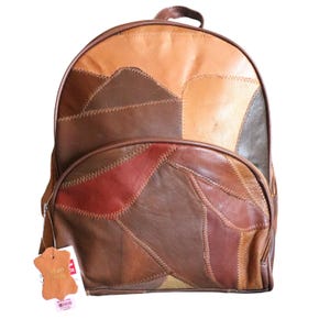 May include: A patchwork leather backpack in shades of brown, tan, and red. The backpack features a top handle, a front zippered pocket, and a main compartment zipper. The design is made of various leather pieces stitched together, creating a unique pattern.
