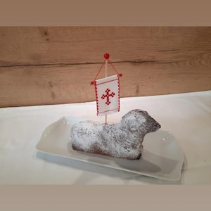 Könnte beinhalten: Ein weißer, lammförmiger Kuchen, der mit Puderzucker verziert ist und mit einer kleinen weißen Flagge mit einem roten Kreuz gekrönt ist.