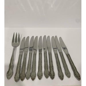 May include: A set of stainless steel silverware, including a fork and several knives. The handles have a decorative, ornate design. The silverware is arranged on a white surface.