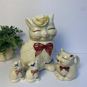 May include: A ceramic cat-shaped cookie jar with a matching creamer and two salt and pepper shakers. The off-white pieces feature hand-painted details including red bows, blue accents, and yellow hats, with closed eyes and whiskers on each feline figure.