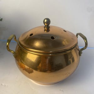 May include: A polished copper pot with a rounded body, two side handles, and a matching lid featuring a small spherical knob and several circular ventilation holes. The metallic surface reflects light, showing a warm, golden-orange hue throughout.