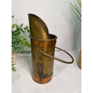 May include: A cylindrical brass and copper coal scuttle featuring a tall, angled scoop insert, a sturdy metal handle, and a small rectangular glass viewing window on the side. The metal surface shows a weathered, aged patina with dark spots.