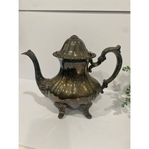 May include: Antique silver-plated teapot with a curved spout, ornate handle, and lid. The teapot has a bulbous body and sits on three small feet. The metal has a tarnished appearance, giving it a vintage look. A classic piece for tea service.