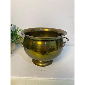 May include: A round, hammered brass bowl featuring a wide, smooth rim and two small, curved side handles. The bowl rests on a circular pedestal base and displays a polished, metallic gold-toned finish with subtle variations in surface texture.