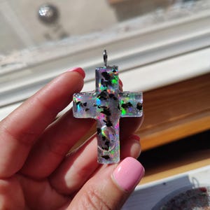 May include: A clear resin cross pendant with iridescent glitter and black flecks. The cross has a silver-colored bail for hanging. The pendant is held up by a hand with pink nail polish.