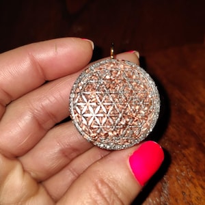 Könnte beinhalten: Ein runder Anhänger mit einem geometrischen Design in Silber und Roségold. Der Anhänger wird in einer Hand mit rosafarbenem Nagellack gehalten.