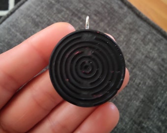 Könnte beinhalten: Ein kreisförmiger schwarzer Anhänger mit einem konzentrischen Labyrinth-Muster und einem silberfarbenen Metallring an der Oberseite. Die Oberfläche des Anhängers ist matt und weist feine rote Sprenkel in der schwarzen Materialstruktur auf.
