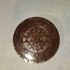 Puede incluir: Un colgante de metal de color cobre con un diseño geométrico detallado en el centro. El diseño está rodeado por un borde decorativo.