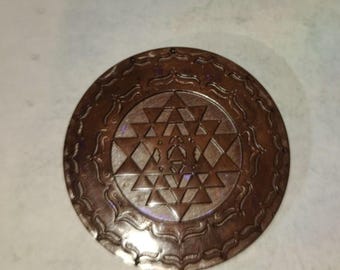 Peut inclure: Un pendentif en métal de couleur cuivre avec un motif géométrique détaillé au centre. Le motif est entouré d'une bordure décorative.