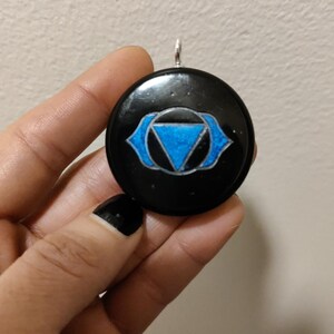 May include: Black pendant with a blue third eye symbol, also known as the Ajna chakra, painted on it.
