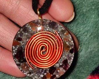 May include: A circular pendant necklace with a black cord. The pendant has a copper wire spiral design over a stone composite of grey, purple, and orange tones. The pendant is held by a gold-coloured bail.