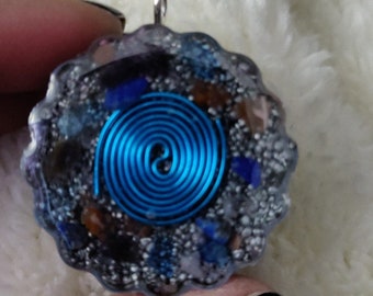 May include: A circular pendant featuring a blue spiral design. The pendant is made of clear resin, encasing blue, brown, and silver-colored stones. It has a silver loop for a chain, suitable for a necklace.