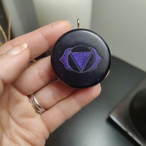 May include: A round, black pendant with a purple third eye chakra symbol. The pendant has a gold bail for hanging on a necklace.