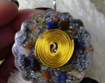 May include: A circular pendant with a silver bail. The pendant has a gold spiral at its centre, encircled by a mix of small, colourful stones and clear resin. The stones include blue, brown, and yellow.