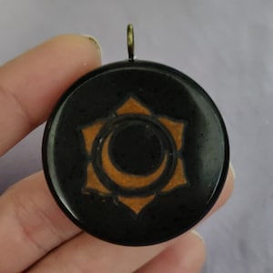 May include: A round black pendant with a gold-colored star and crescent moon design. The pendant has a small loop at the top for a chain. The design is centered on the pendant.