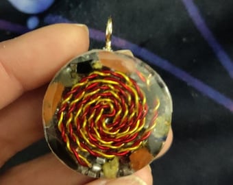 May include: A round pendant with a spiral design. The pendant features a clear resin encasing a red and gold wire spiral, surrounded by small stones and metal pieces. A gold-colored loop is attached at the top for hanging.