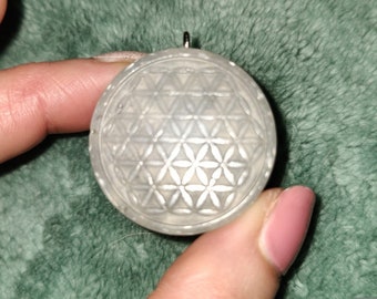 May include: A white, round pendant with a detailed, geometric design resembling a flower of life. The pendant has a silver loop at the top for attaching a chain.